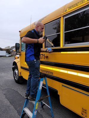 Man on ladder applying sealant to school bus window. Yellow bus, gray sky.