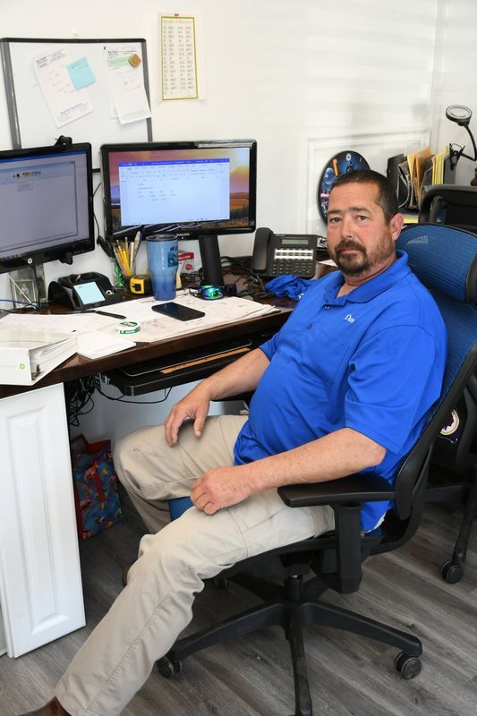 Man in blue shirt seated at a desk with two monitors and a phone. Beige pants, office setting.