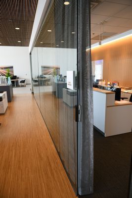 Glass wall separating an office space with wooden floors, visible desks and chairs, and a mesh curtain.