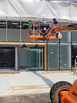 Construction workers on a lift, installing a structure, in front of a building with glass doors and windows.