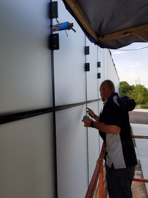 Man on a lift applying sealant to a gray building's exterior panels. Black trim and brackets are visible.