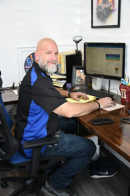 Man with beard at a desk, typing on a computer. He is in an office, wearing a blue and black shirt.