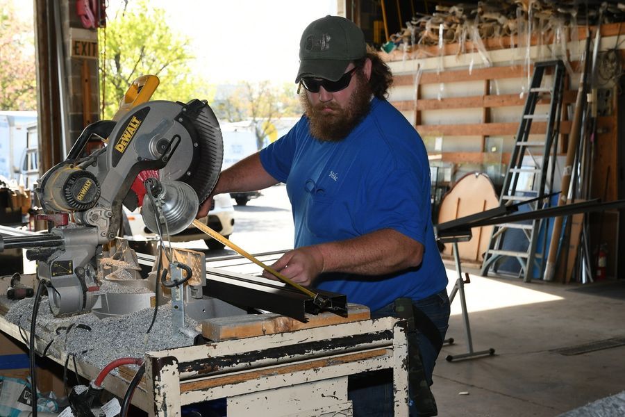 Man in blue shirt using a miter saw in a workshop, measuring a black beam.