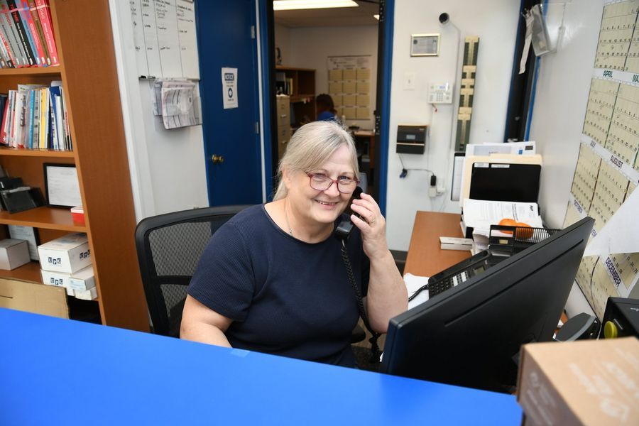 Woman at desk, smiling, on the phone. Blue counter, office setting, shelves with books.