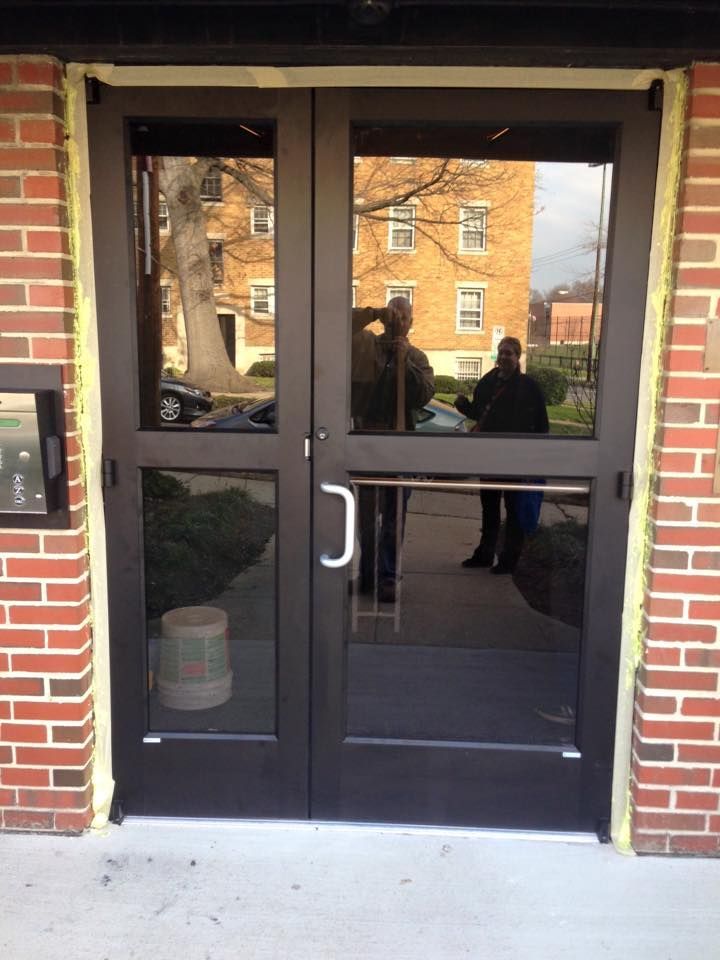 Double dark-framed glass doors reflecting a building and people. Brick exterior, light-colored trim.