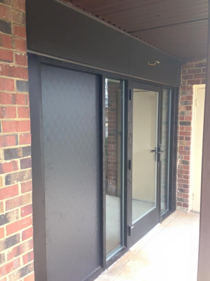 Exterior view of dark-framed glass doors and a solid black panel. The doors are set in a brick building.