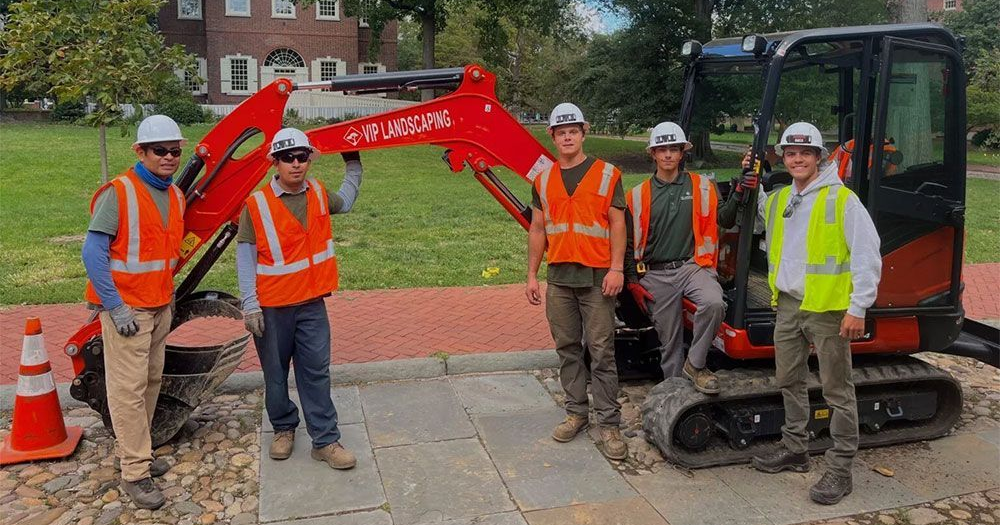 Five construction workers in safety vests pose by a small red excavator on a brick and stone walkway.