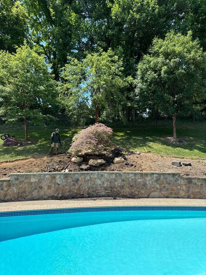 A person working on a hillside garden near a pool; green trees and blue sky background.