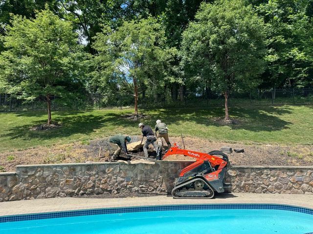 Workers using a small excavator to remove vegetation from a hillside near a pool.
