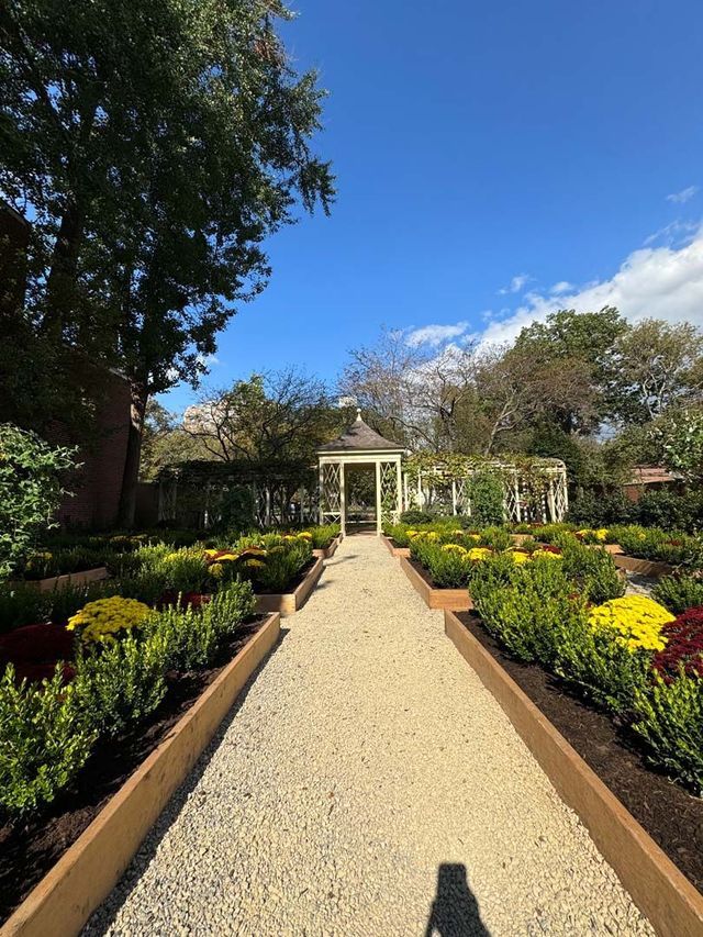 A gravel path leads to a gazebo through a manicured garden with colorful flowers and green hedges under a blue sky.