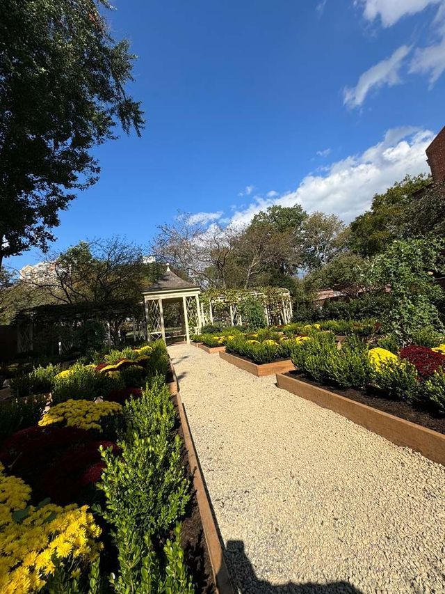 Gravel path through a vibrant garden with a white gazebo under a bright blue sky.