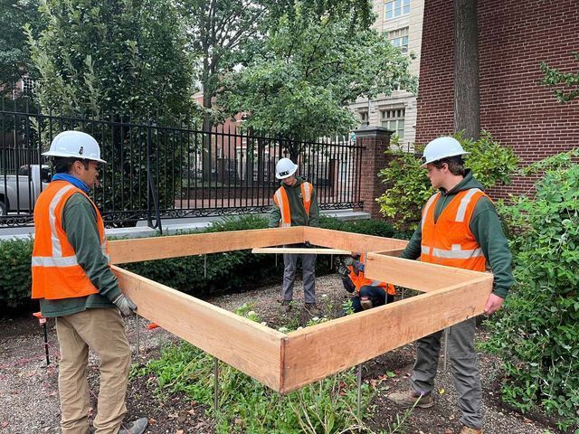 Three workers in safety vests and hard hats lift a square wooden frame outdoors, near a building.