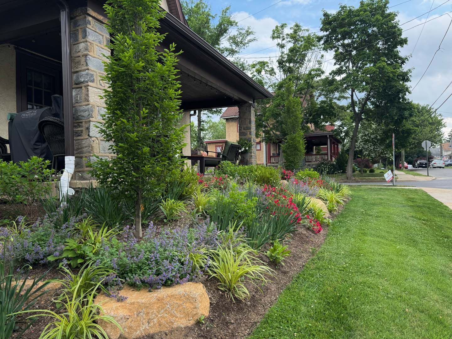 A colorful landscaped garden bed in front of a stone house with green grass and trees.