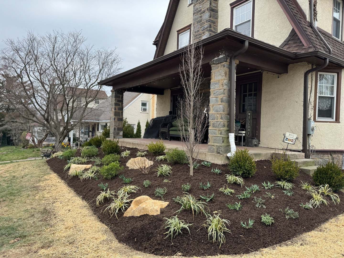 Landscaped front yard with brown mulch, shrubs, and a stone-columned porch in front of a house.