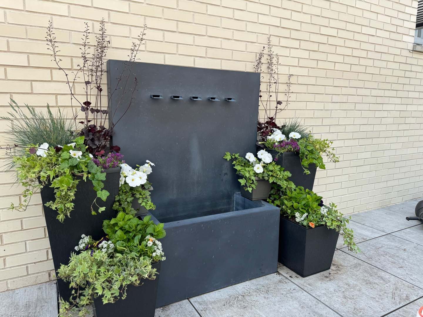 Water fountain with planters on rooftop. Gray fountain, black planters with flowers against brick wall.