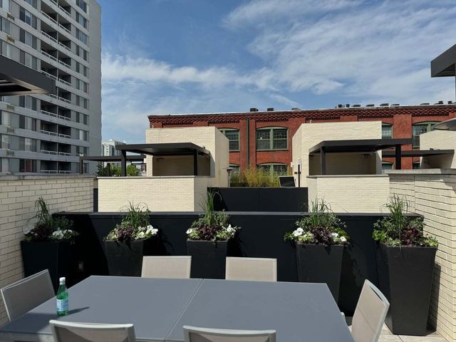 Rooftop patio with seating, black planters with flowers, against a building backdrop under a blue sky.