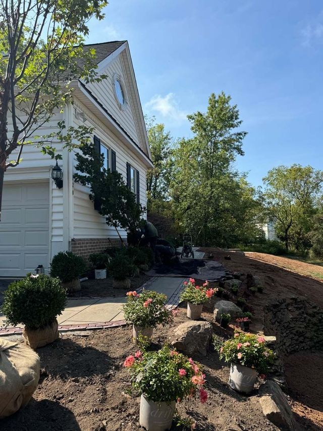 Exterior of a house during landscaping. Person working with plants and rocks; pink flowers, green bushes.