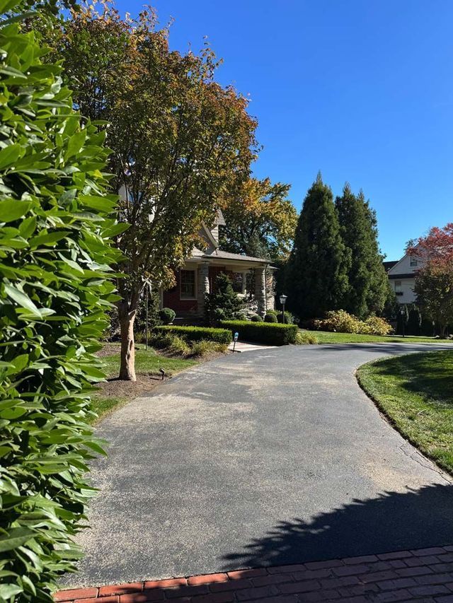 Asphalt driveway leading to a house with a porch and surrounding greenery under a blue sky.