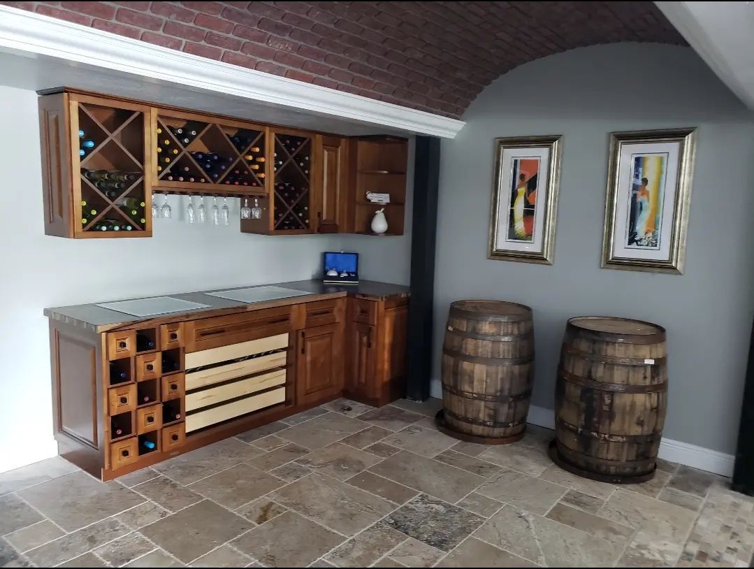 Rustic indoor bar with wooden cabinets, wine racks, two barrels, and framed art on a stone floor