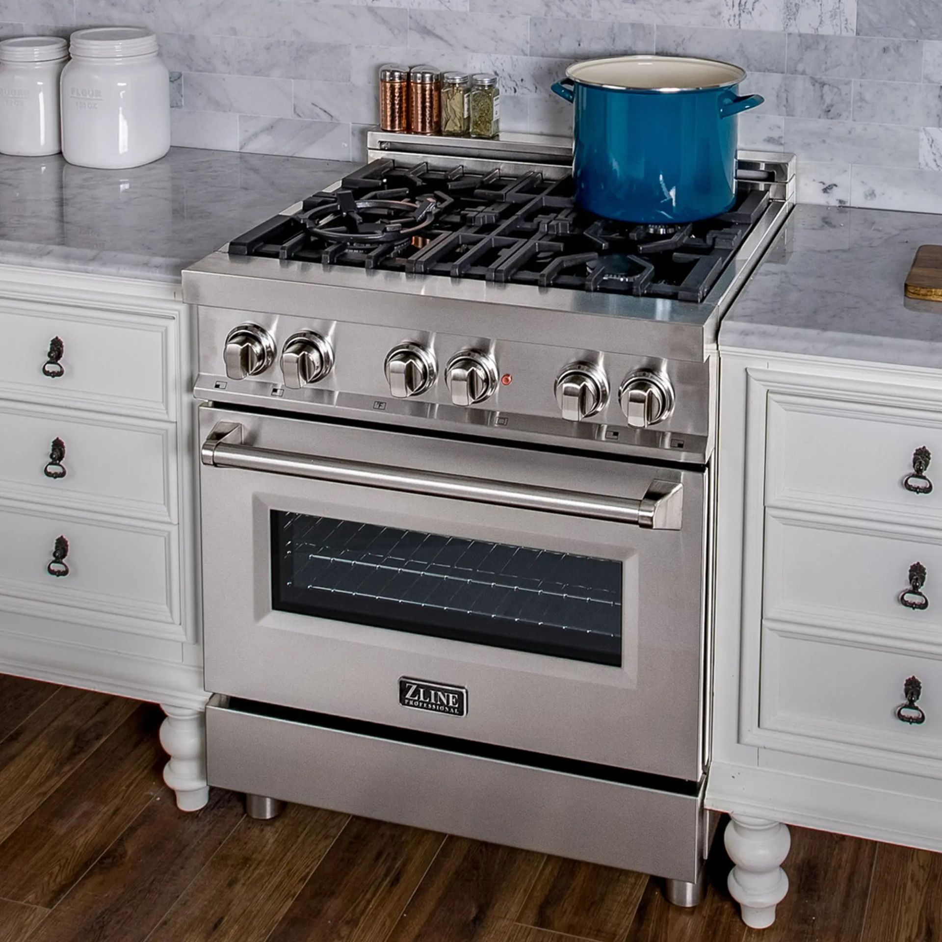 Stainless steel gas range with blue pot on burners in a white kitchen with marble backsplash