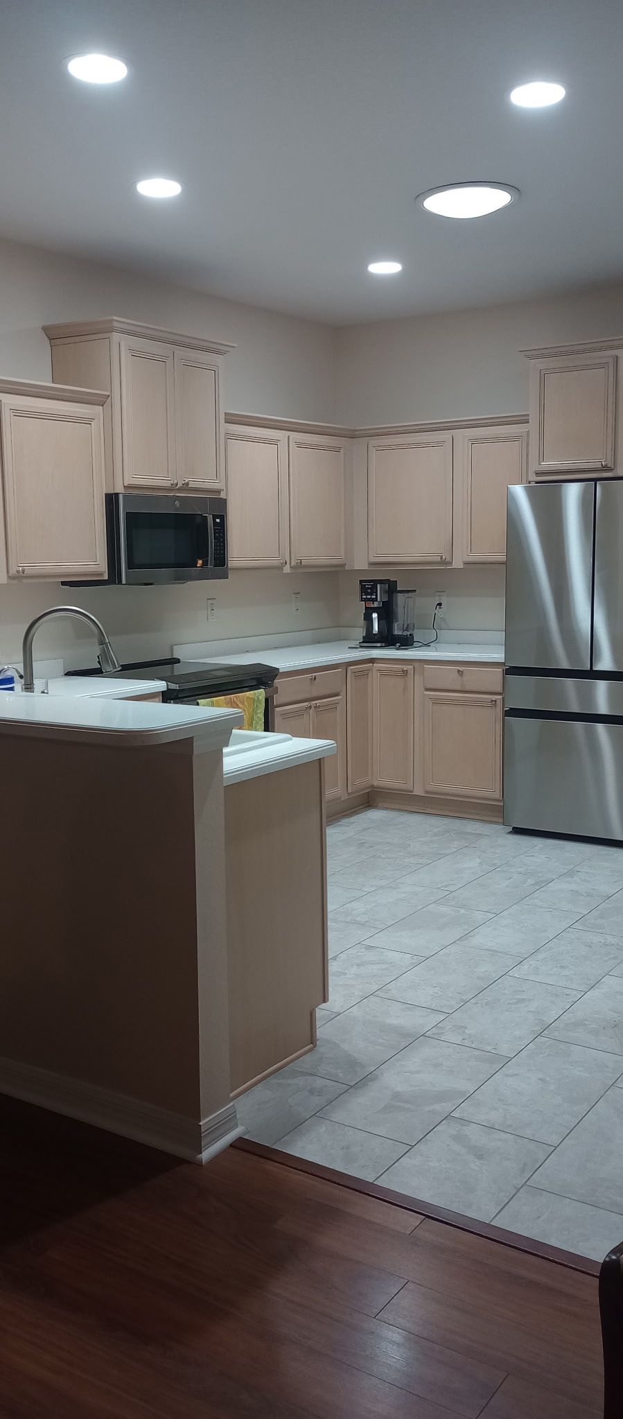 Empty modern kitchen with beige cabinets, stainless steel fridge, and bright recessed ceiling lights