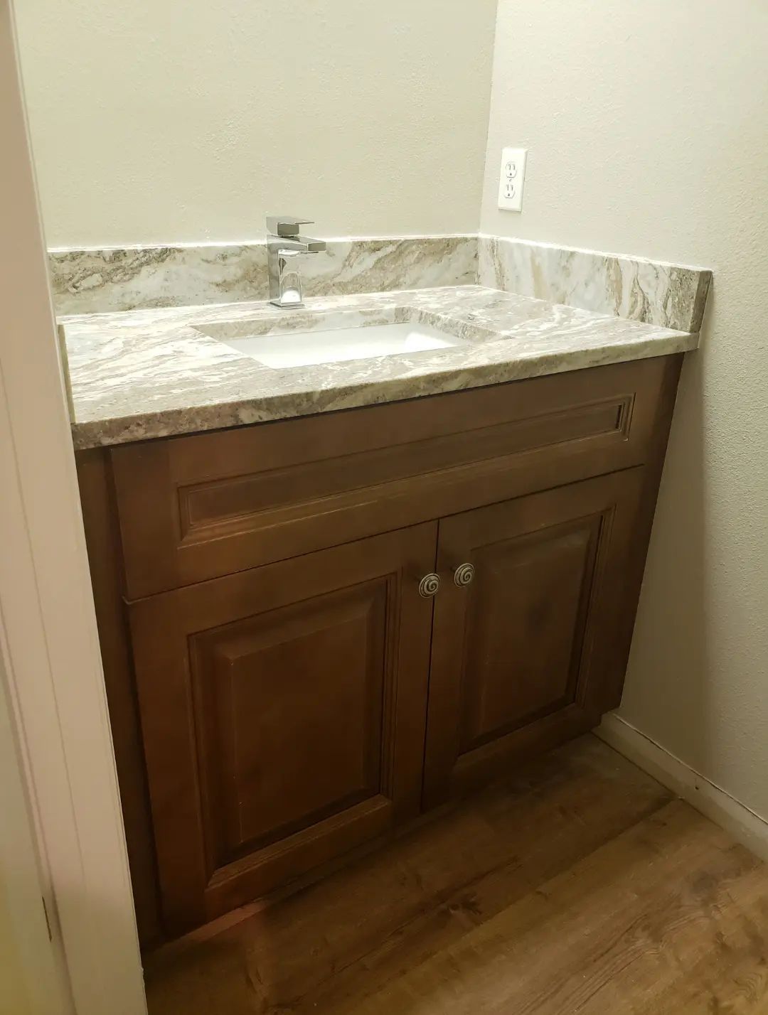 Small bathroom vanity with wood cabinet, marble countertop, and sink against a beige wall.