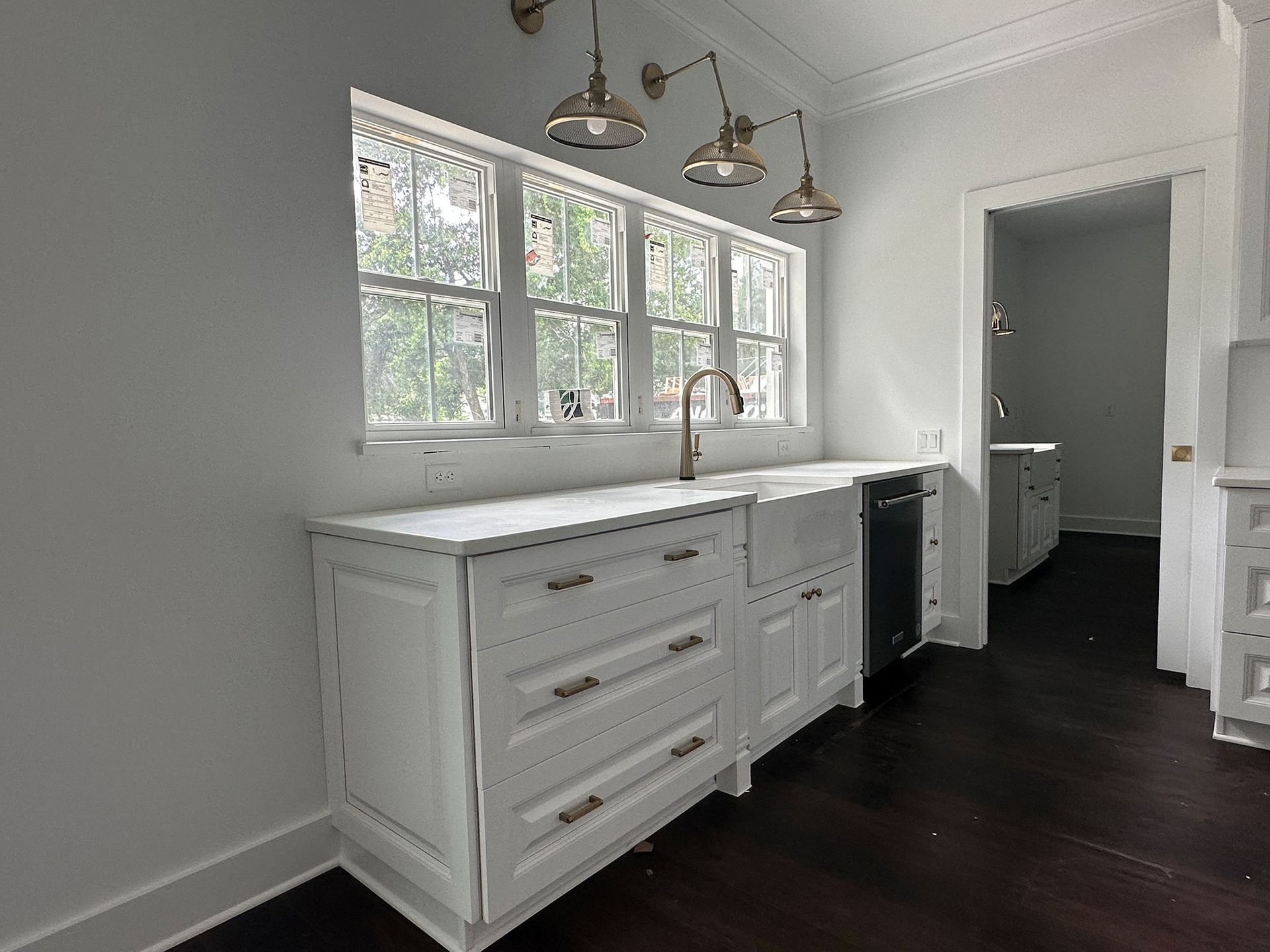 Bright white kitchen with window, sink, and dark wood floor leading to a small bathroom doorway