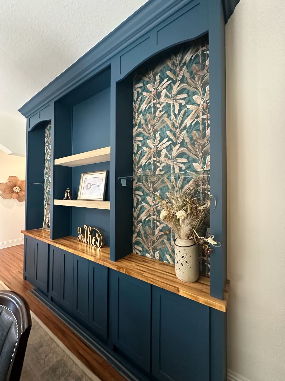 Blue built-in cabinet with floral wallpaper panels, wood shelf, and decorative vase in a living room.