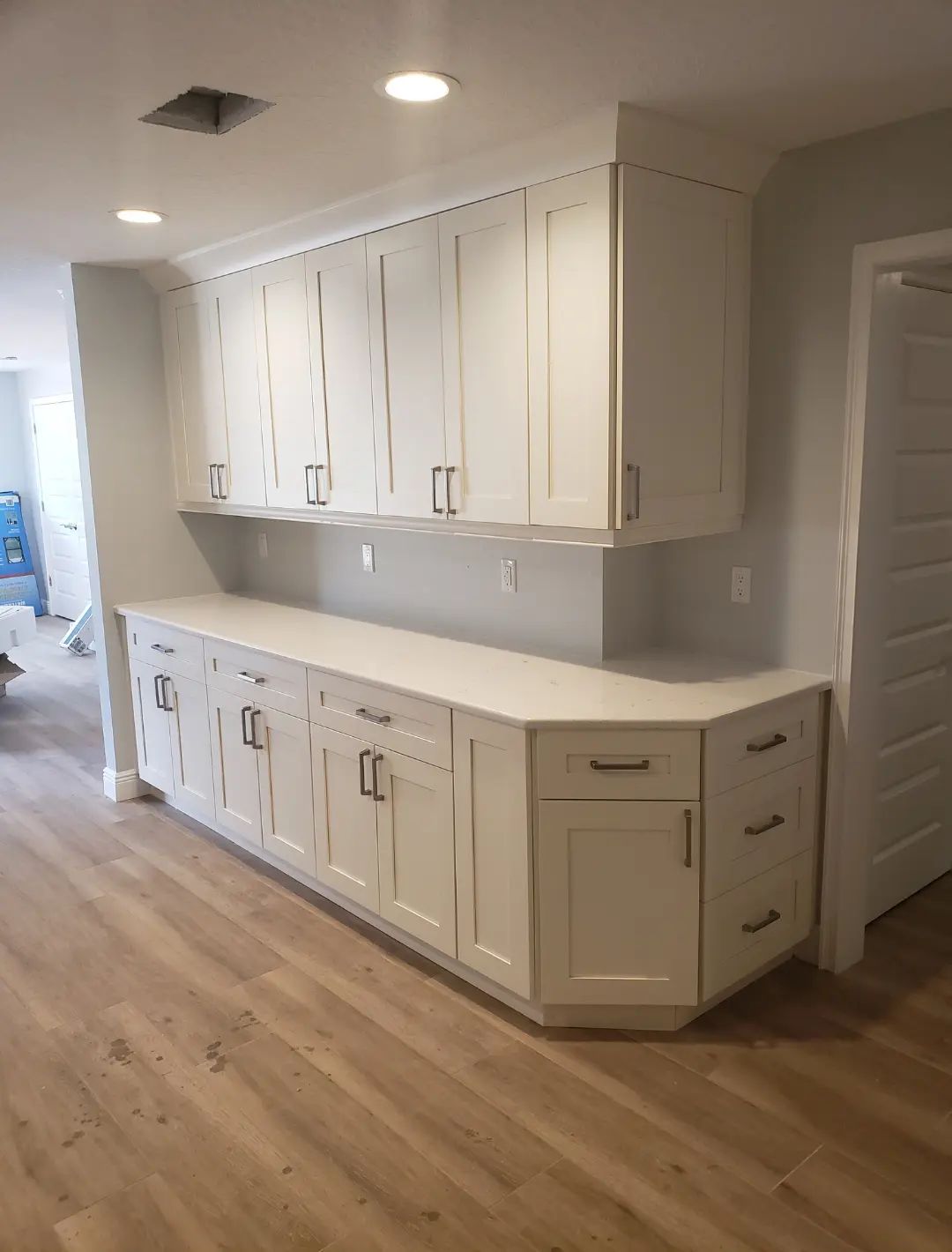 White built-in kitchen cabinets and countertop along a hallway, with light wood floors and recessed ceiling lights.