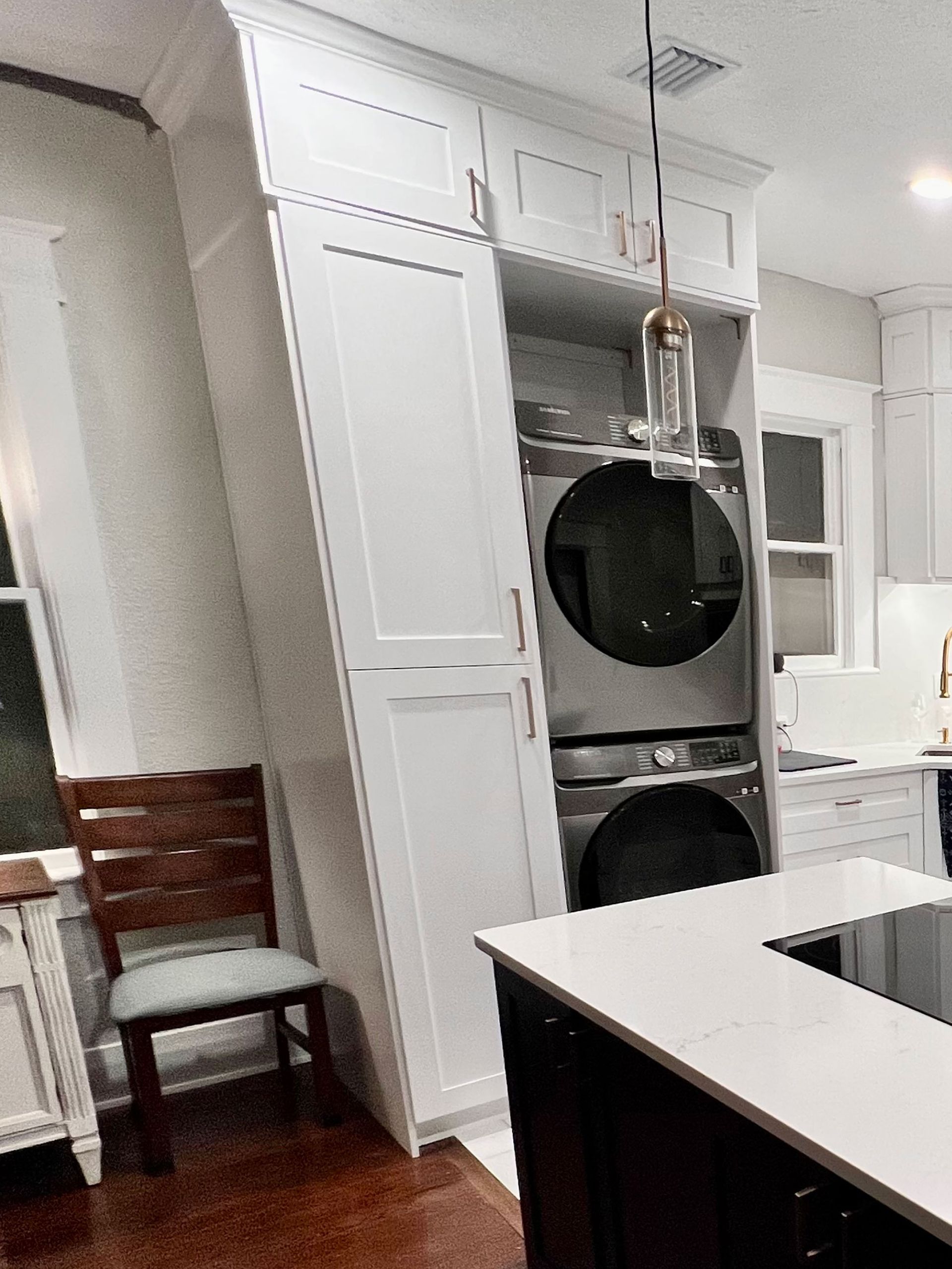 White laundry room with stacked washer and dryer beside a kitchen island and chair.