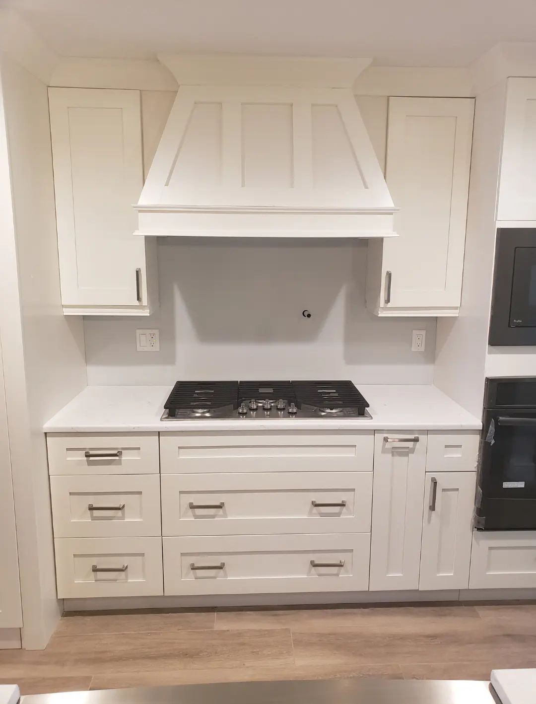 White kitchen with built-in cabinetry, central range hood, black cooktop, and light wood floor.