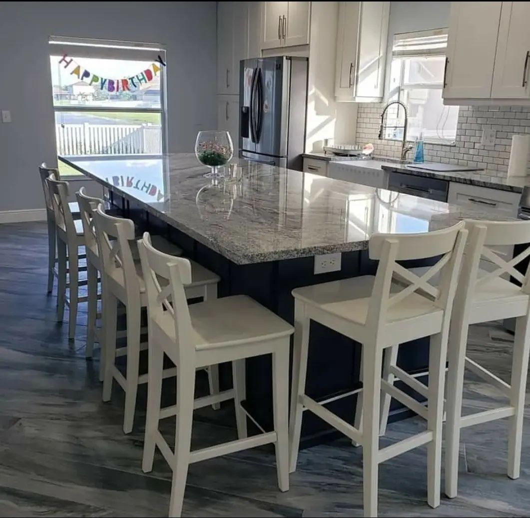 Bright kitchen with a large granite island, white barstools, stainless fridge, and window seat.