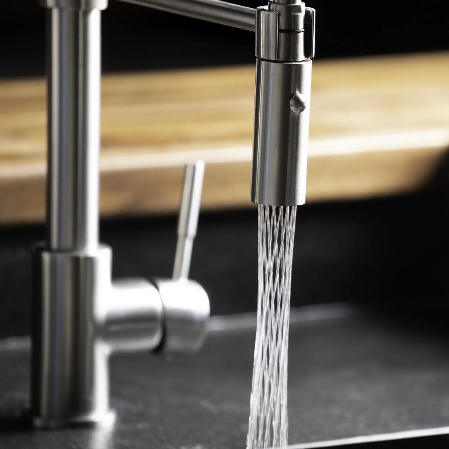 Stainless steel kitchen faucet running water into a sink, with a blurred wooden background.