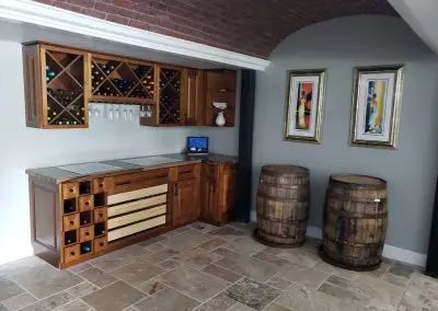 Wine cellar bar with wood cabinetry, countertop, barrel decor, and framed art on a stone-tile floor.