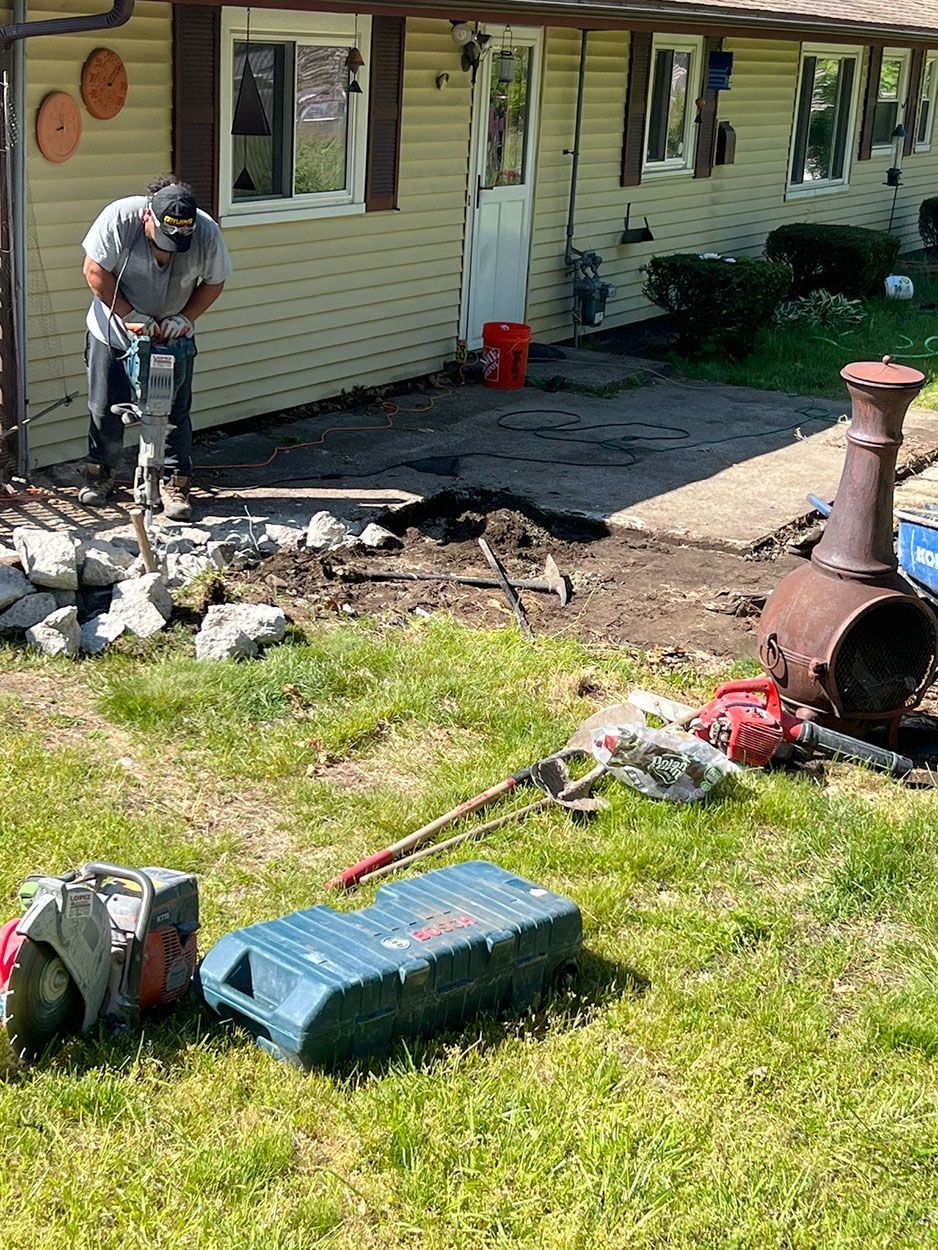 A man is using a jack hammer to break the concrete slab in front of a house