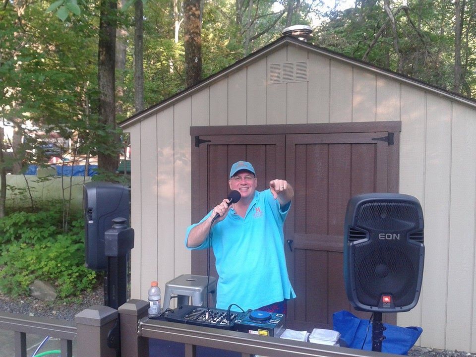 A man in a blue shirt is standing in front of a shed holding a microphone.