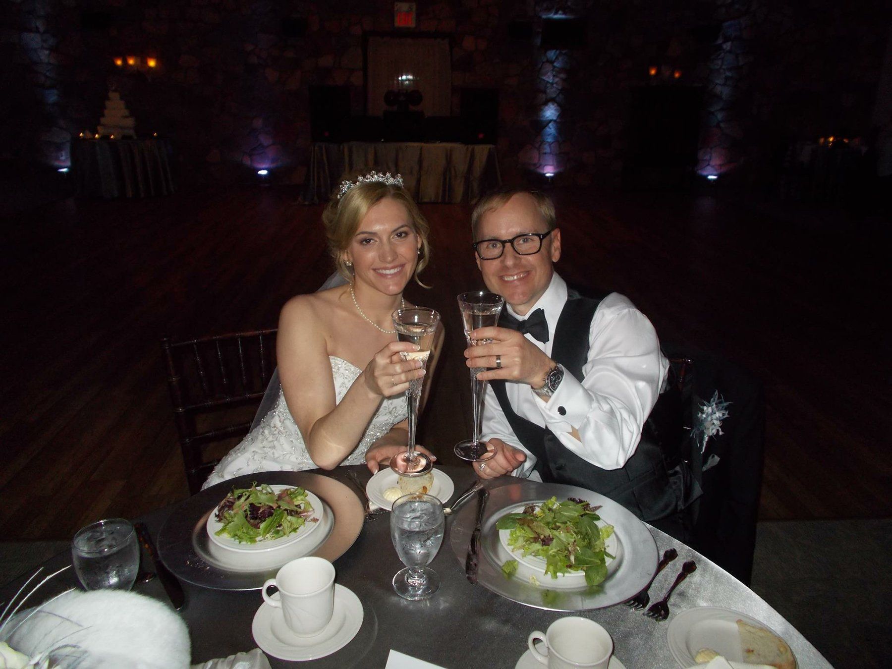A bride and groom are sitting at a table with plates of food and glasses of wine