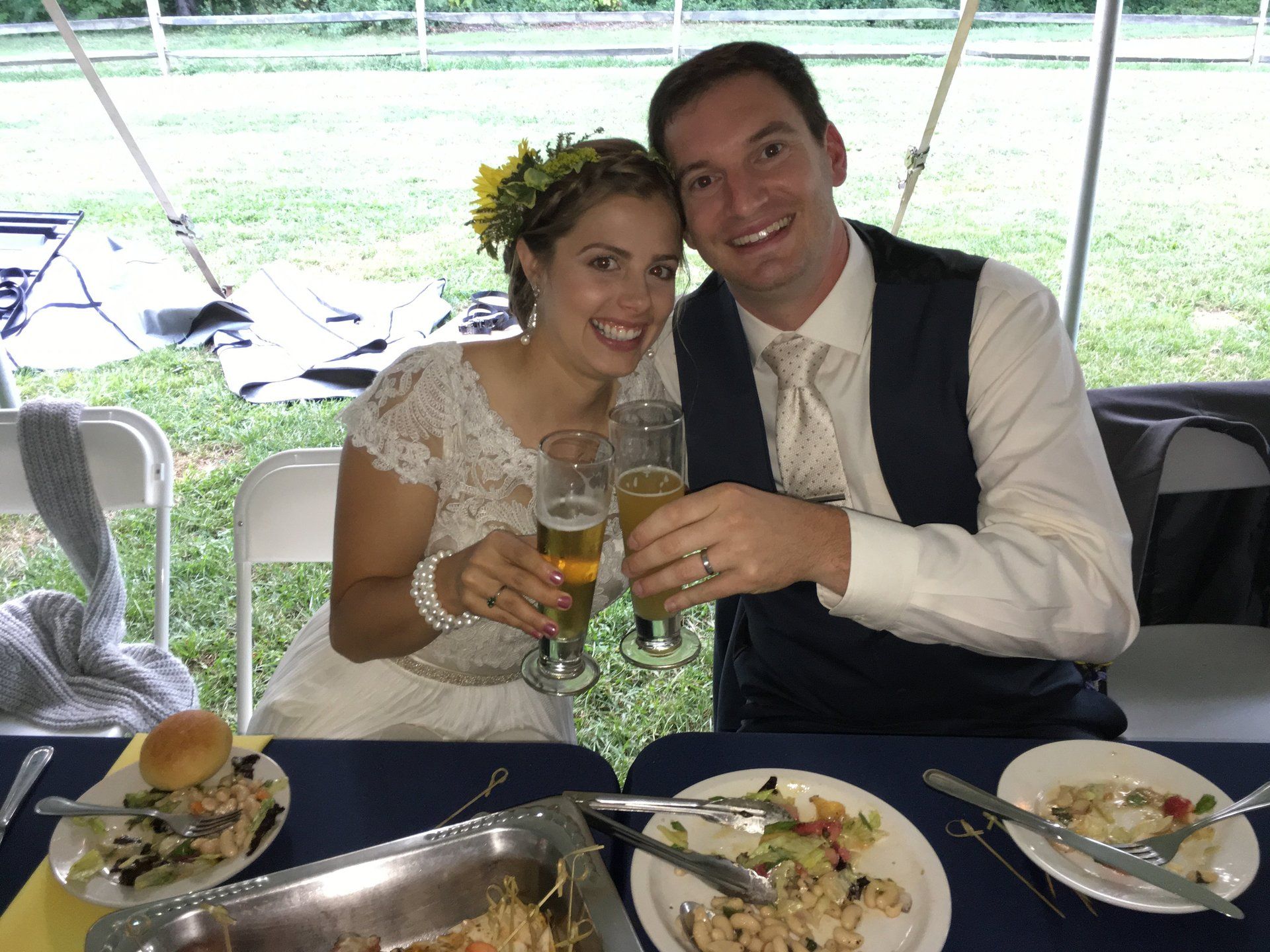A bride and groom are sitting at a table with plates of food and glasses of beer.