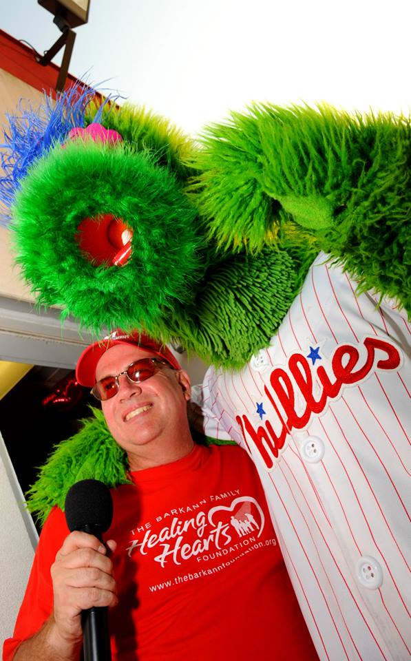 A man is holding a microphone next to a phillies mascot.
