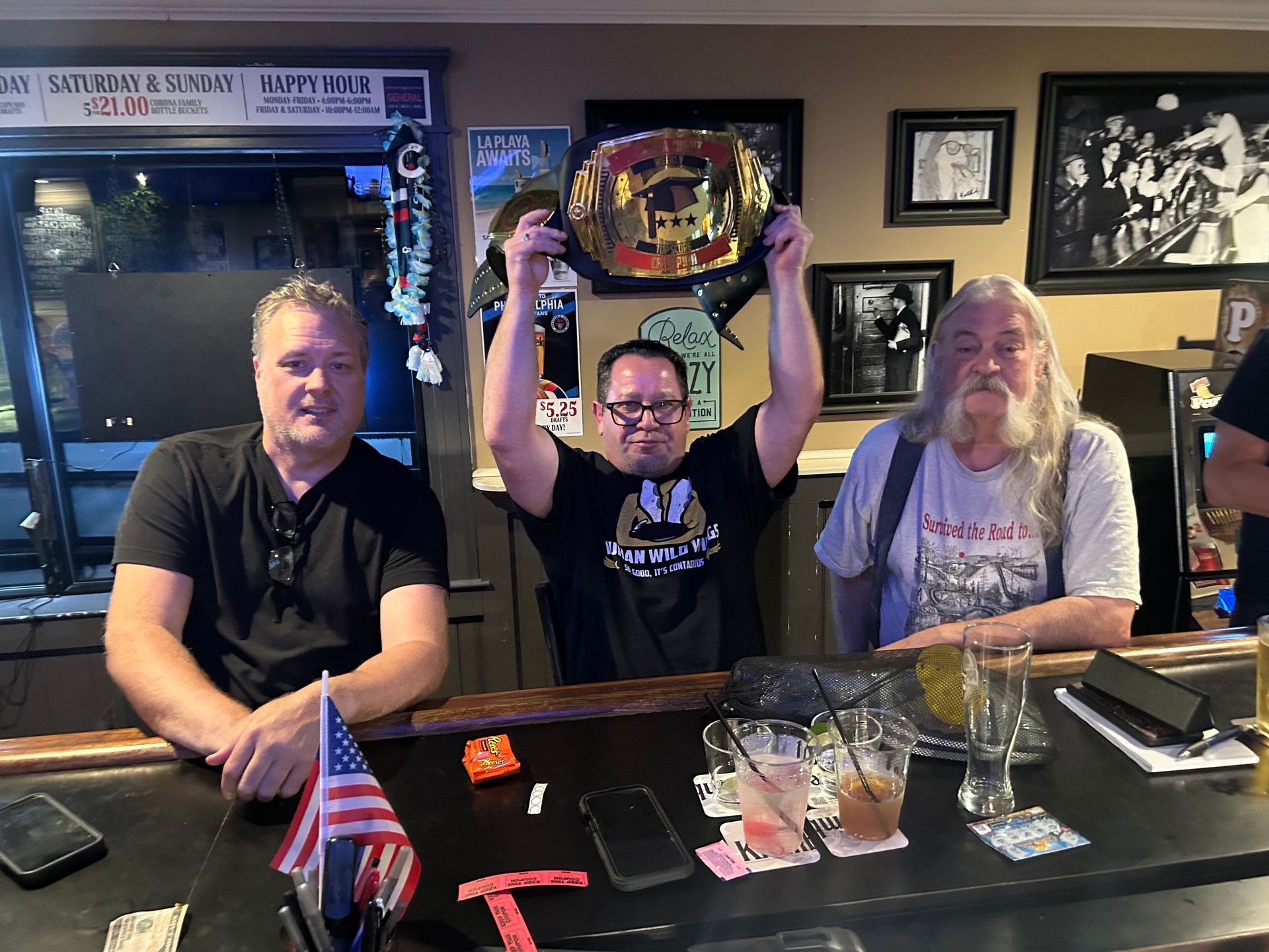 Three people at a bar, one holding a championship belt overhead, drinks and flags on the counter.