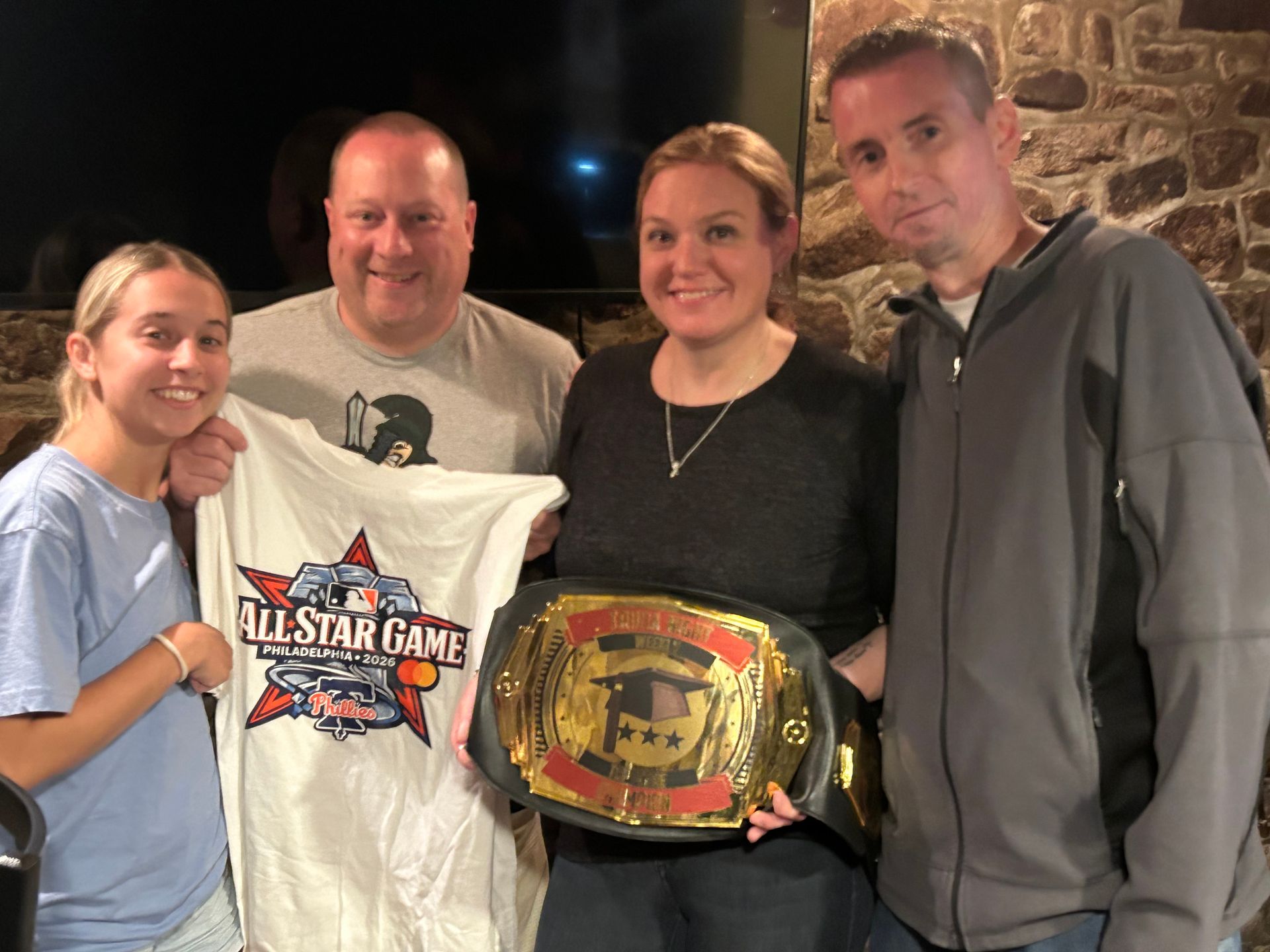 Four people posing indoors, two holding wrestling championship belts and one holding a T-shirt.
