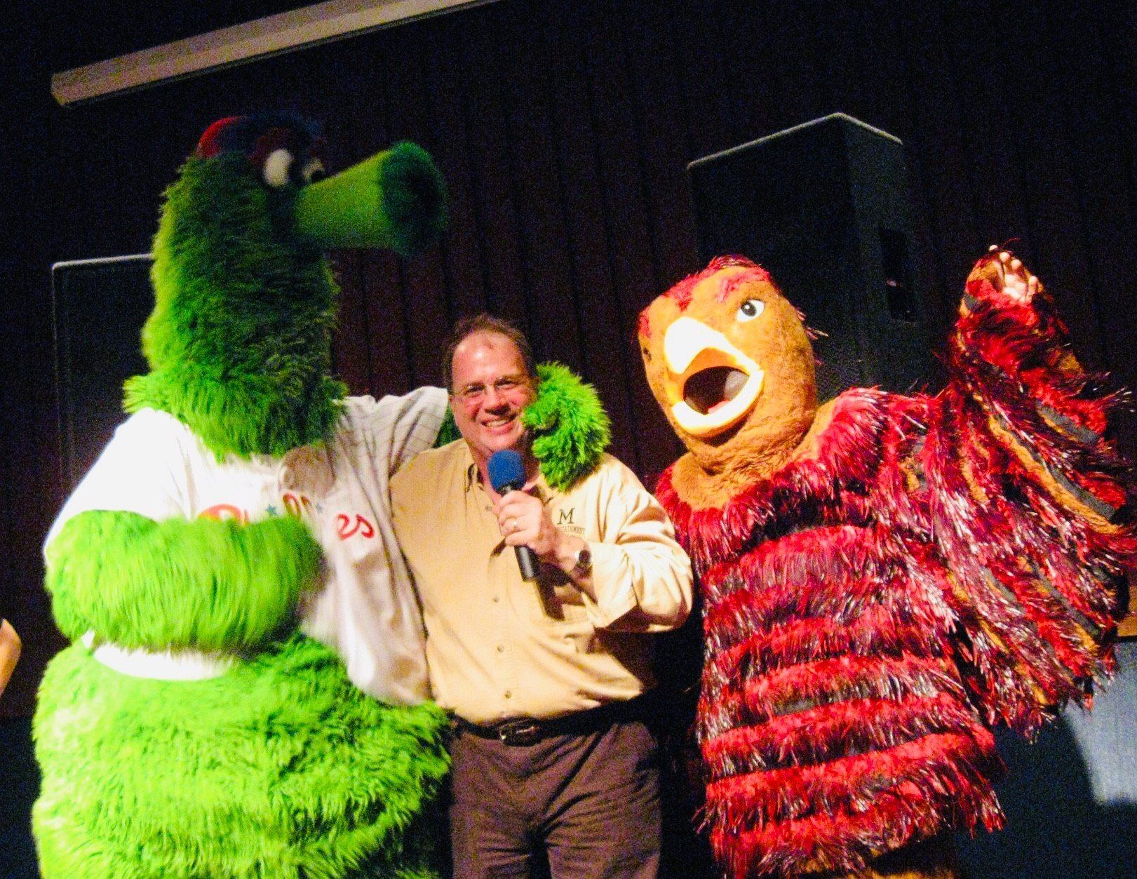 A man with a microphone stands between two mascots