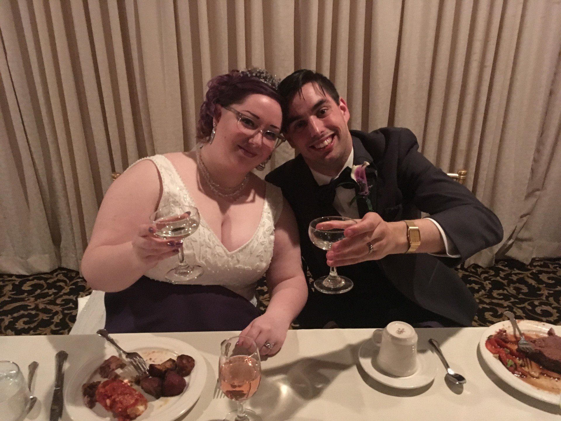 A bride and groom are sitting at a table with plates of food and glasses of wine.