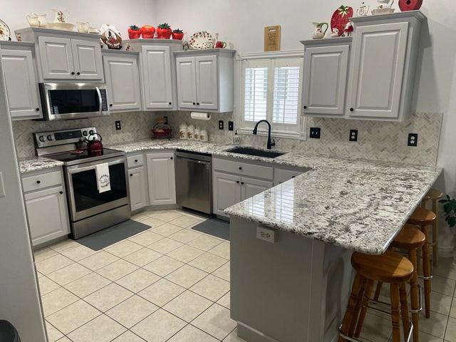 Bright kitchen with gray cabinets, white tile floor, granite counters, and a large island with stools.