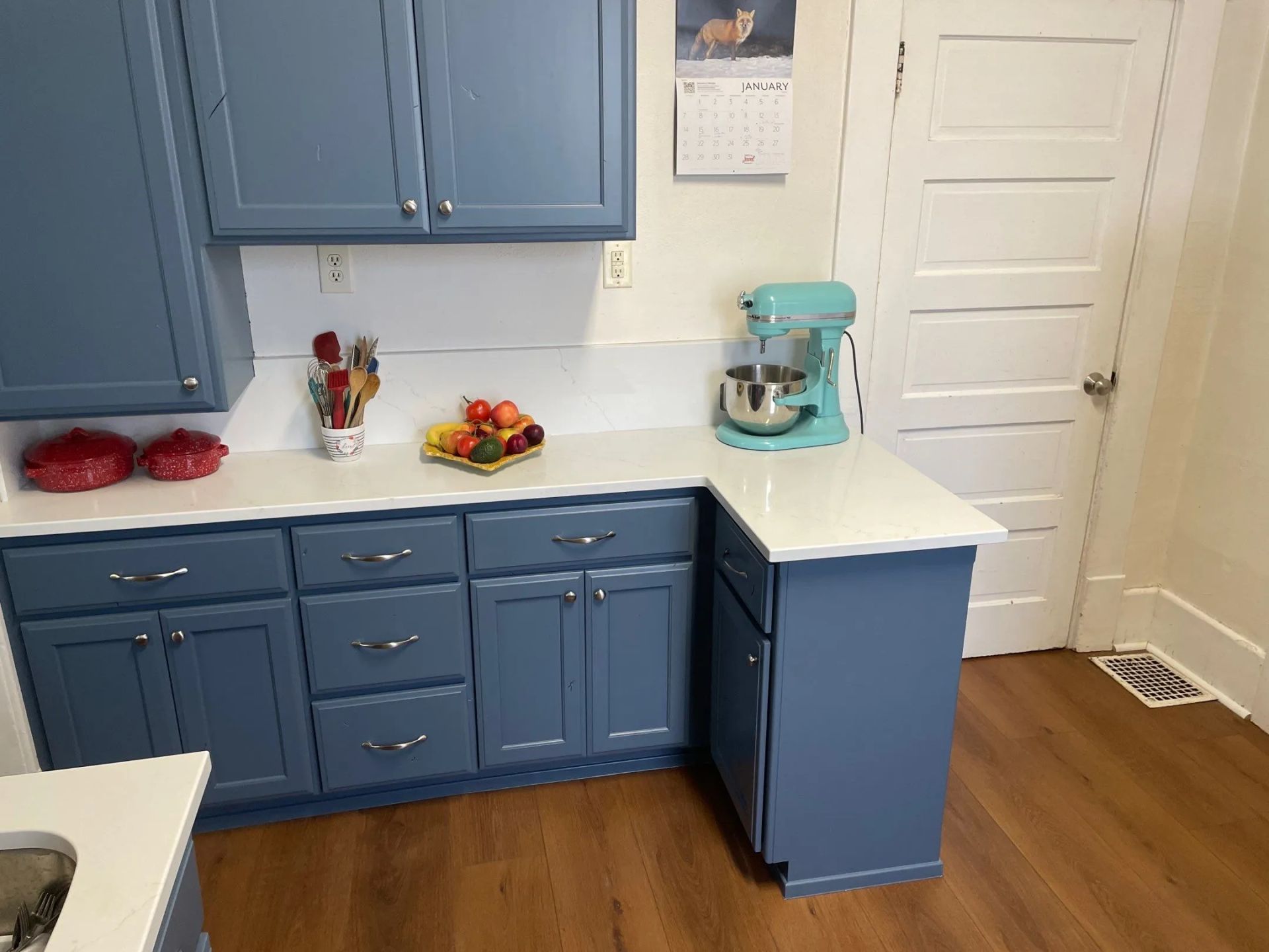 Blue kitchen cabinets and white countertops with a teal stand mixer on the counter and fruit bowl near a door