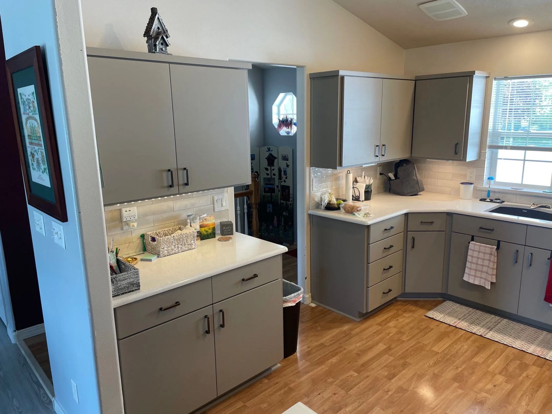 Bright kitchen with gray cabinets, white countertops, wood floor, and a window over the sink.