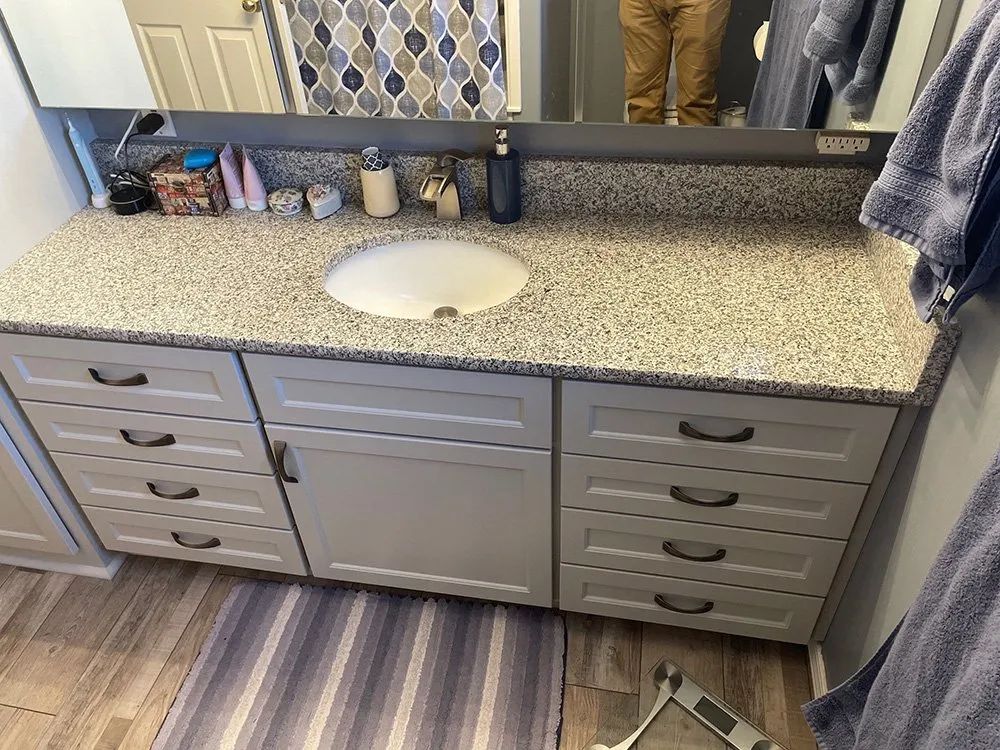 Gray bathroom vanity with speckled countertop, white sink, drawers, mirror, and striped rug.