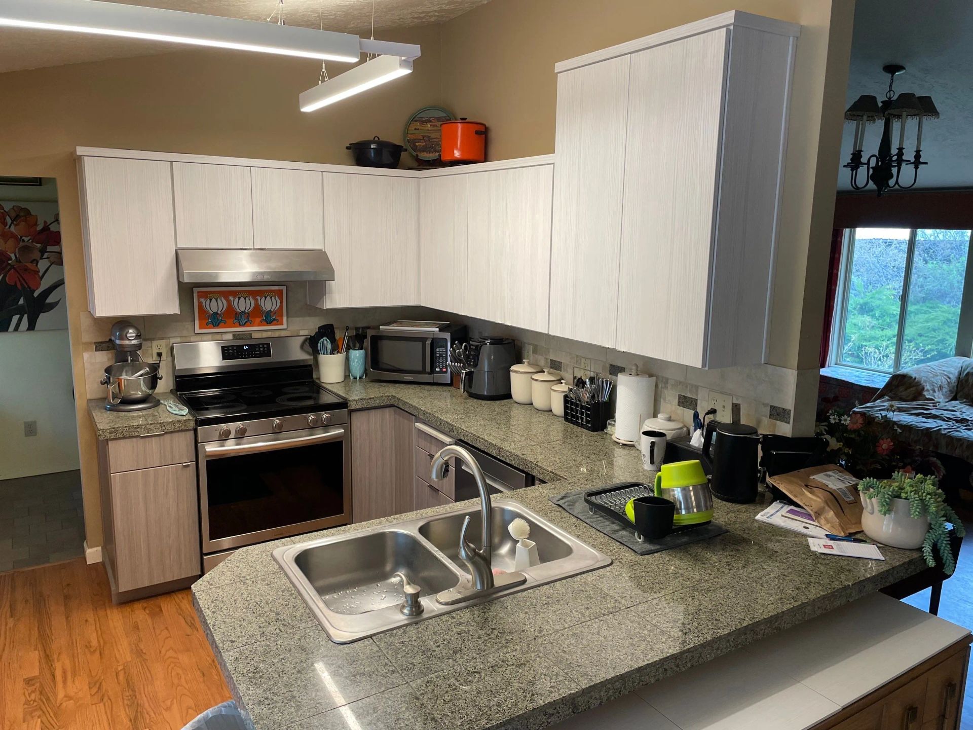 Bright kitchen with white cabinets, granite island, sink, stove, and window overlooking greenery.
