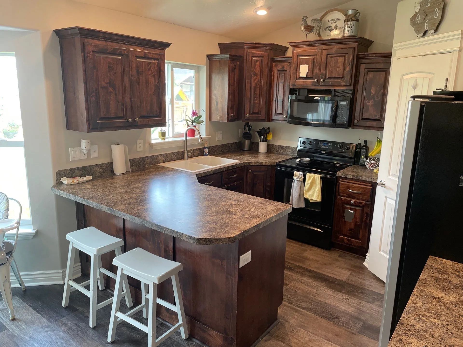 Warm kitchen with dark wood cabinets, granite island, and two white stools by a window