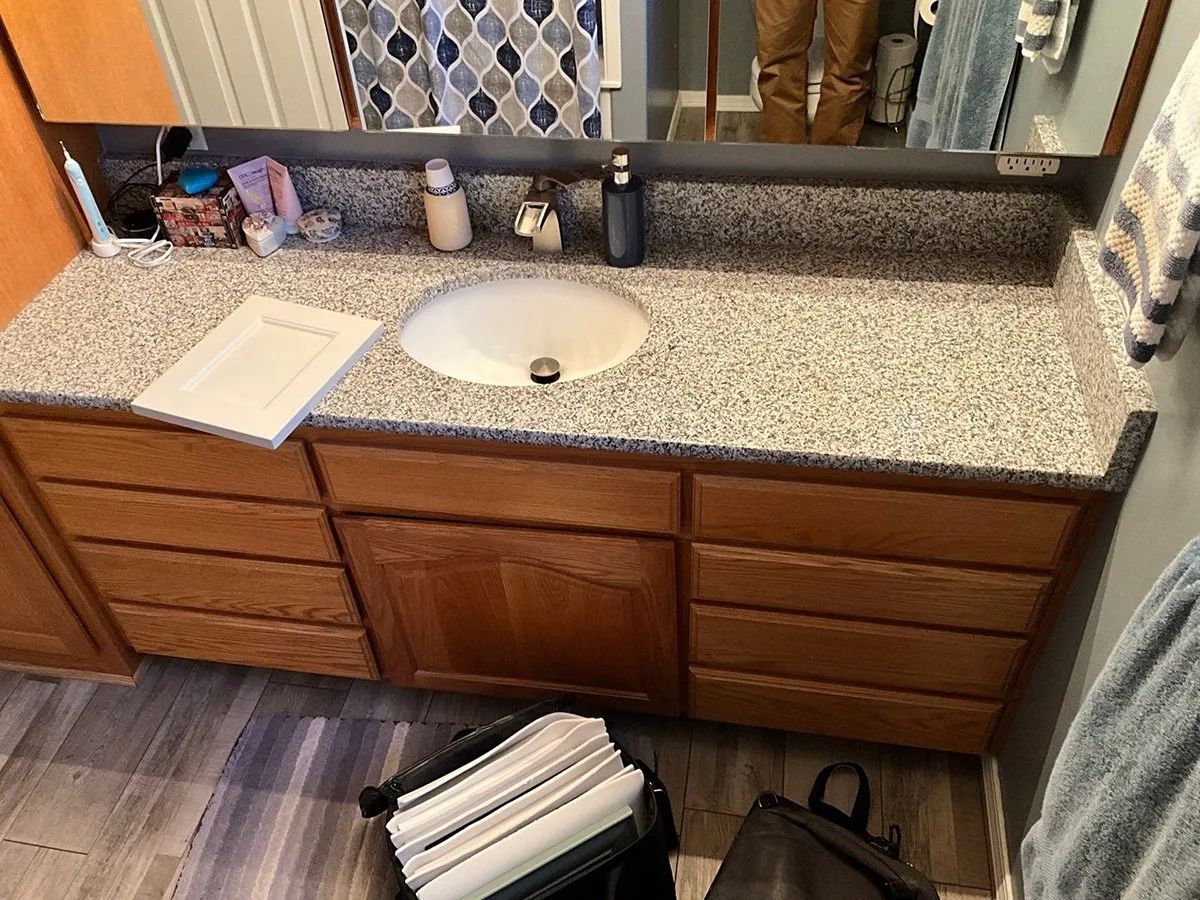 Bathroom vanity with speckled countertop, sink, mirror, and wooden cabinets; open drawer on the left.
